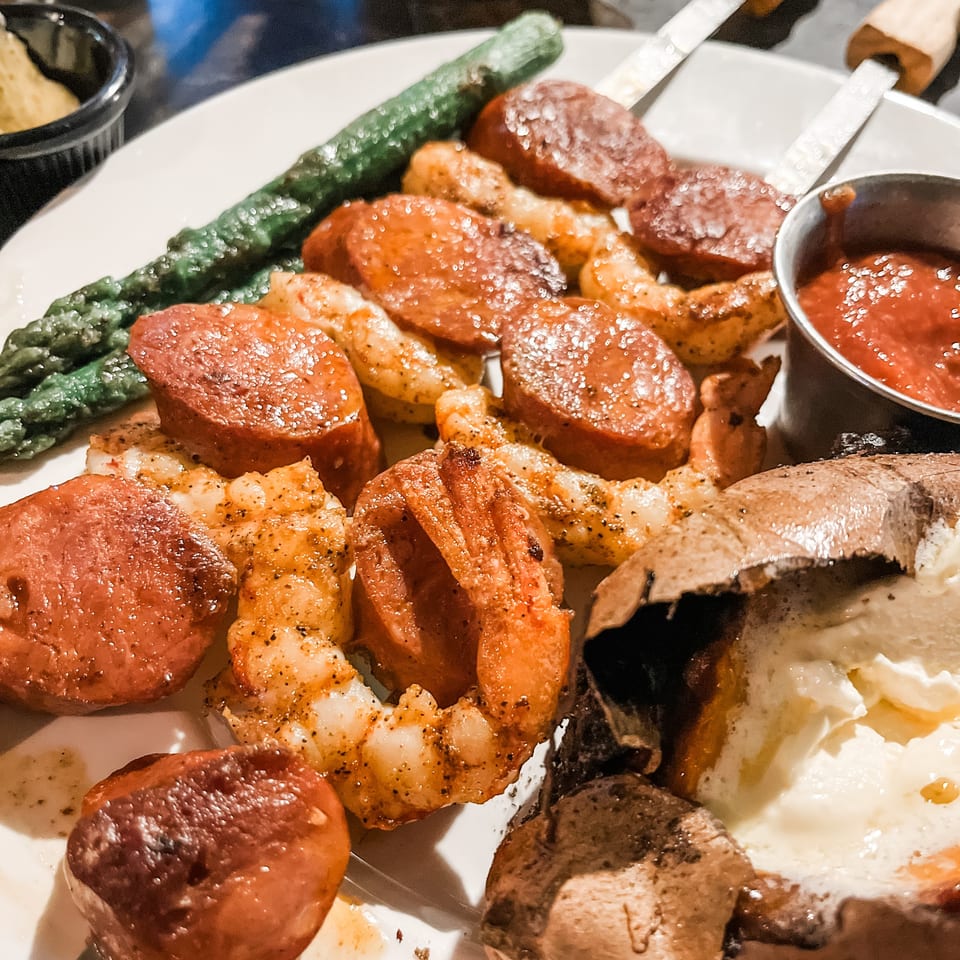 Grilled shrimp and sausage with asparagus, a baked potato, and dipping sauce on a white plate.