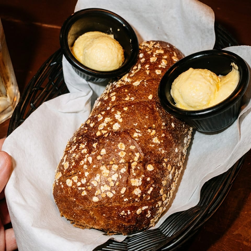 A hand reaches for a basket of dark bread with oats, served with two small cups of butter.